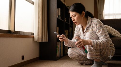 自宅のリビングで、市販の殺虫剤を手に持ち、家具の隙間へ逃げ込む害虫をスマートフォンのライトで照らしながら、困り果てた表情で見つめる日本人女性。精神的な疲弊とプロへの相談を検討する様子。