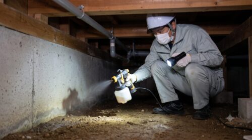 日本の戸建て住宅の床下で、日本人作業員が合成殺虫剤を散布し、シロアリ防除のバリア工法を行っている様子。