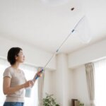 天井のゴキブリに虫取り網を近づけ、凍結スプレーを噴霧する日本人女性。安全な高所駆除の様子。