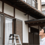 コウモリ被害のある自宅を背景に、ハシゴでは届かない2階の軒先を見上げて困惑する日本人の男性住人。