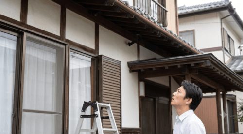 コウモリ被害のある自宅を背景に、ハシゴでは届かない2階の軒先を見上げて困惑する日本人の男性住人。