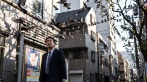 都市部の公園で、多数のドバトが留まる電線や木の下を歩く日本人男性。宝くじのポスターの前を通り、空から何かが落ちてくる様子。