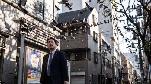 都市部の公園で、多数のドバトが留まる電線や木の下を歩く日本人男性。宝くじのポスターの前を通り、空から何かが落ちてくる様子。