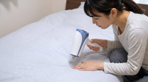 ベッドの上の布団にヘアドライヤーを近づけて熱風を当てるアジア人の女性の様子。