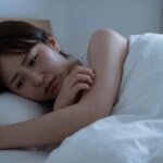寝具の中で腕をかき、ダニ被害に悩む日本人女性の様子