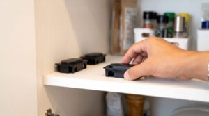 清潔なキッチンの棚に、日本人の男性の手がチャバネゴキブリ用の黒いベイト剤（毒餌剤）を設置している様子。