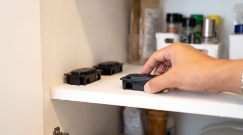 清潔なキッチンの棚に、日本人の男性の手がチャバネゴキブリ用の黒いベイト剤（毒餌剤）を設置している様子。